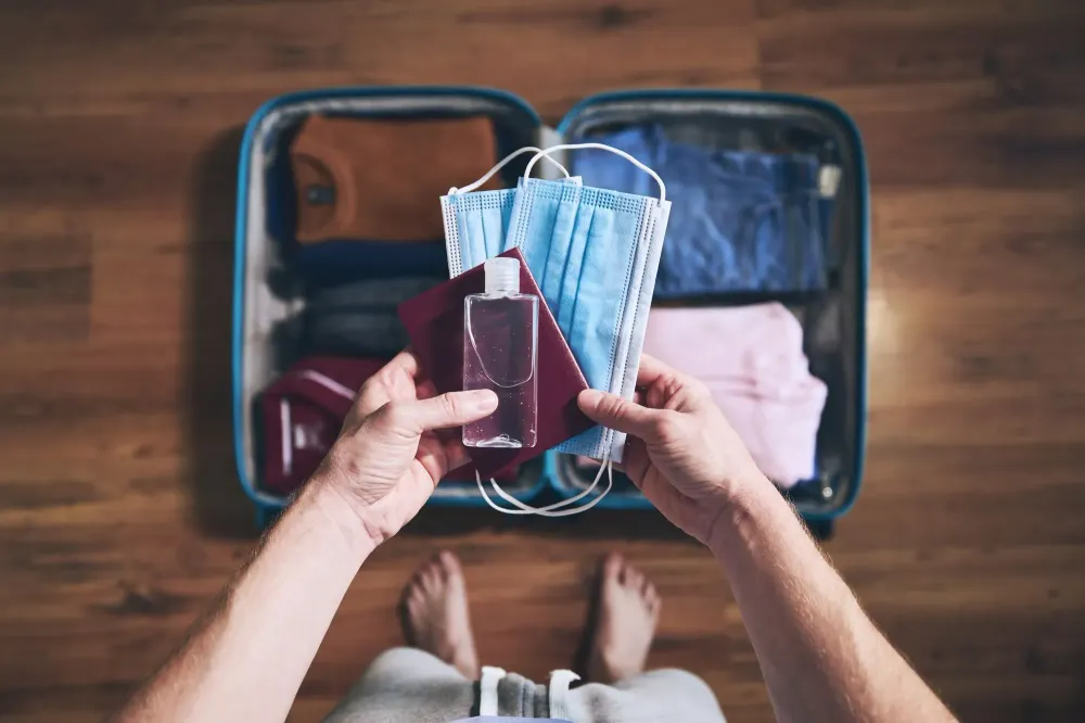Person holding a face mask, passport book, and hand sanitizer over a packed suitcase on the floor
